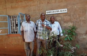 Three Kenyan men standing in front of a building with a sign that says Administration Block